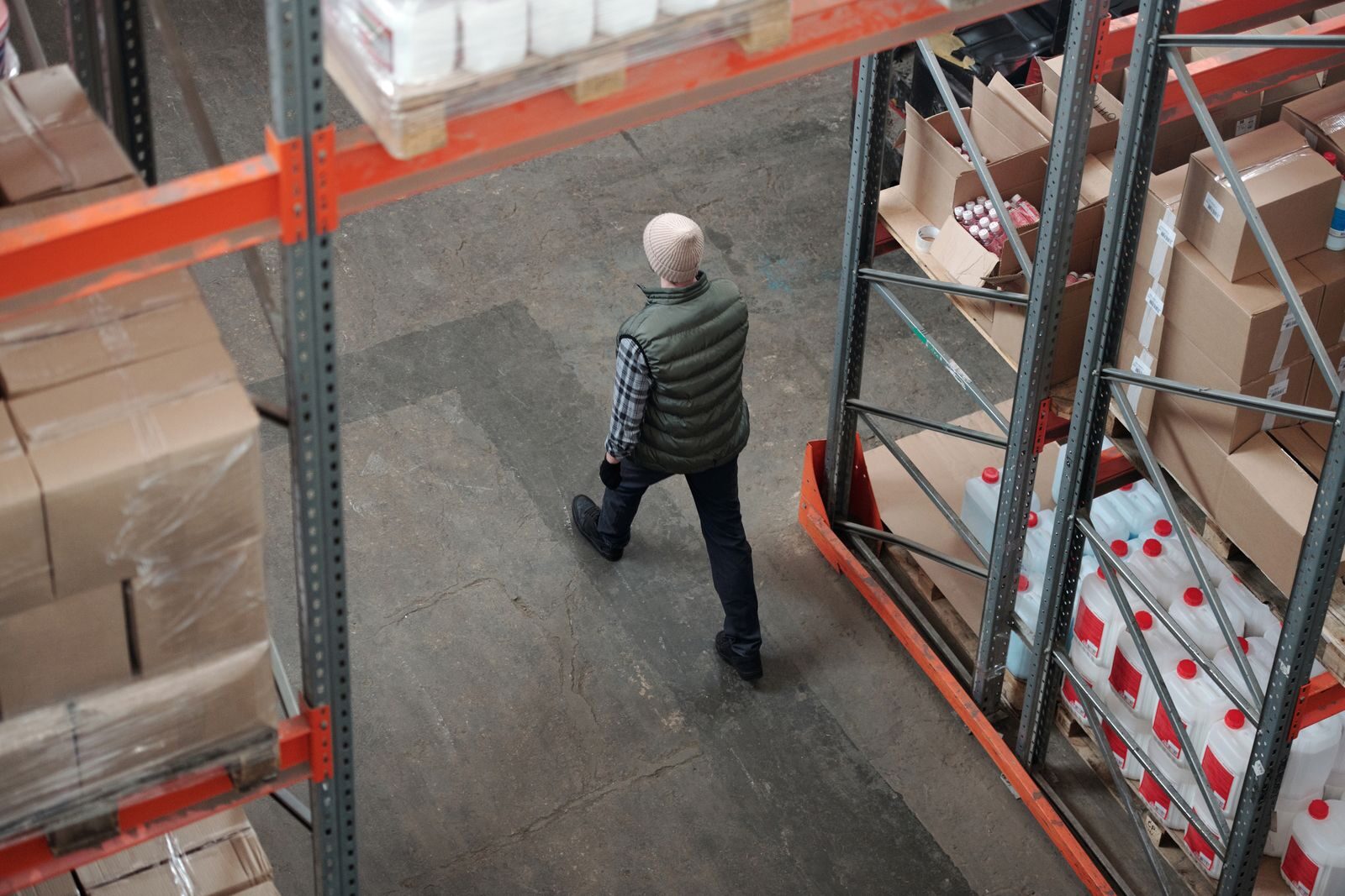 Photo by Tiger Lily Overhead view of a worker walking through a warehouse aisle surrounded by shelves of packages.